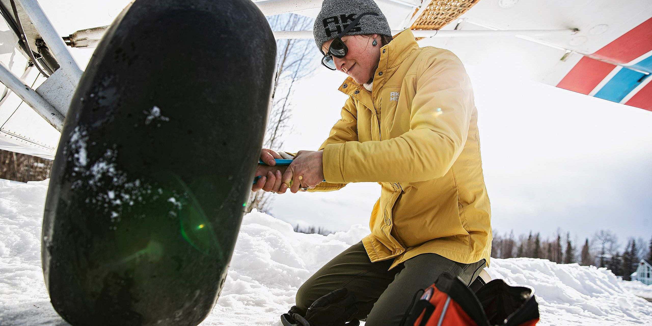 Person in a yellow jacket fixes tire on a bush plane on a snowfield.