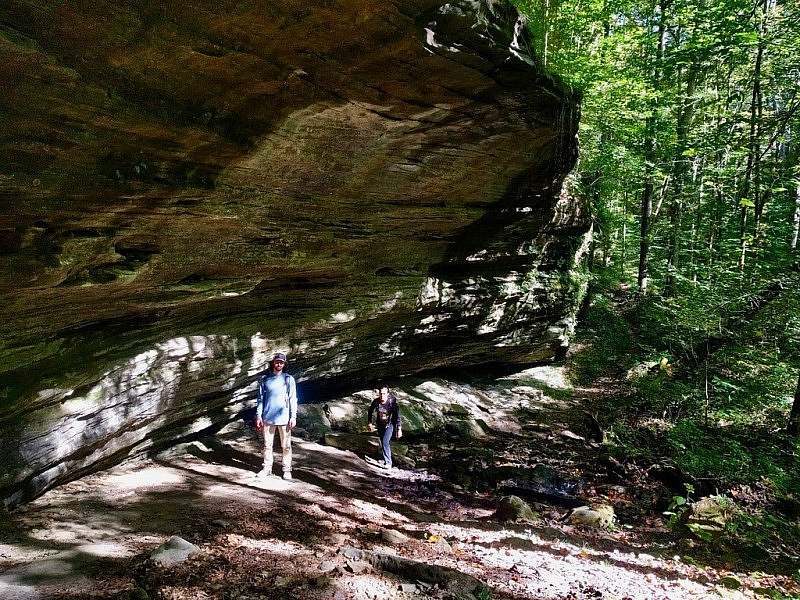 Two hikers stand below an overhanging cliff with a thick green forest beyond the rock formation. 