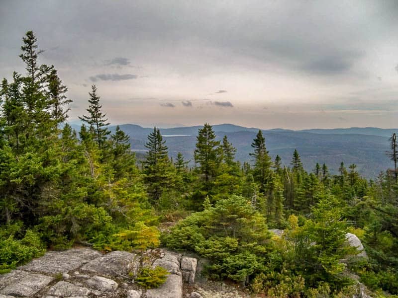 Thick evergreens cover all but the rocks on a hillside sloping down into a forested valley with a few lakes in the distance. 