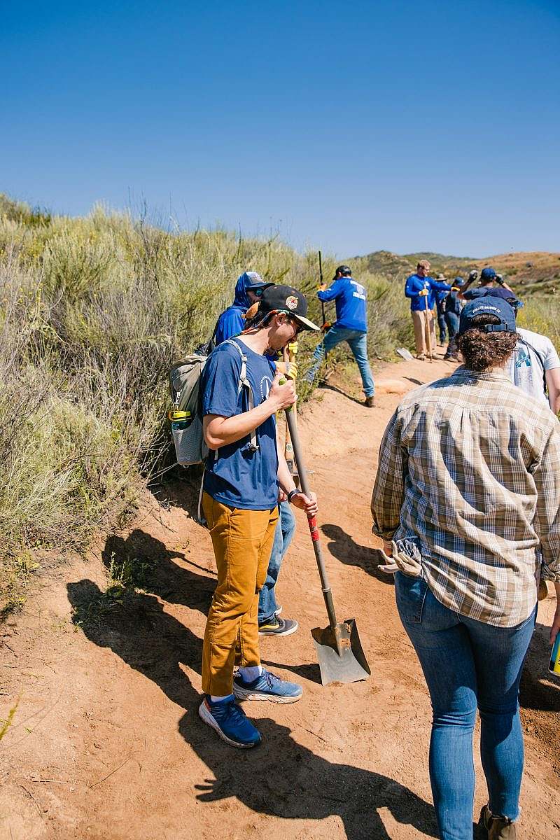 Volunteers are working together on a dirt trail, many are holding trail maintenance tools, like shovels.