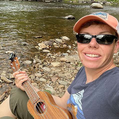 Emily Wallbridge outdoor self portrait along the banks of a river wearing a baseball hat and sunglasses and holding a ukulele.