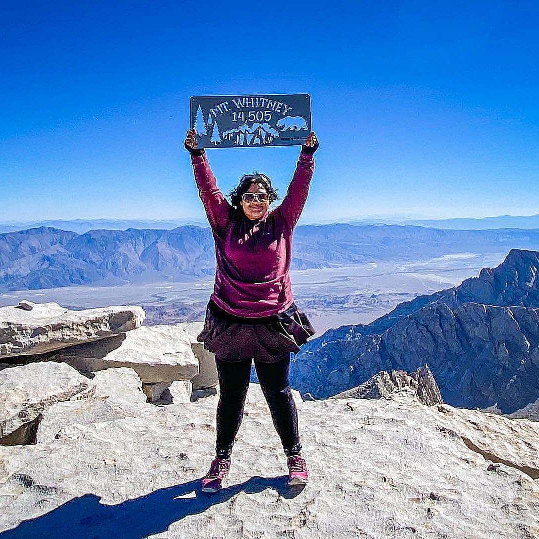 A Hiker stands on top of Mt. Whitney holding a Mt. Whitney sign.