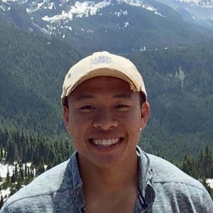 Jeremiah Su smiles for an outdoor portrait in a baseball cap with a forested hillside in the background