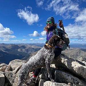 Hike with a dog stands on a rocky summit wearing a warm hatt with blue sky and puffy clouds and mountains in the background