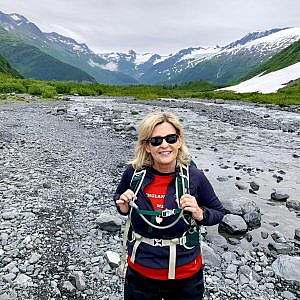 Hiker wearing sunglasses and holding both backpack shoulder straps stands in a rocky valley with snowy mountains in the background