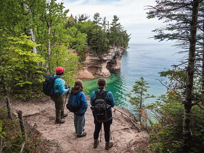 Three hikes gaze at a large body of water from the tree line atop cliffs above the water.