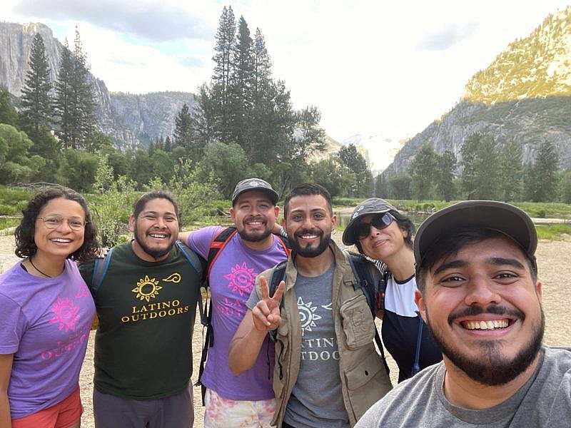 group of hikers wearing Latino Outdoors shirts smile for a selfie photo in a meadow.